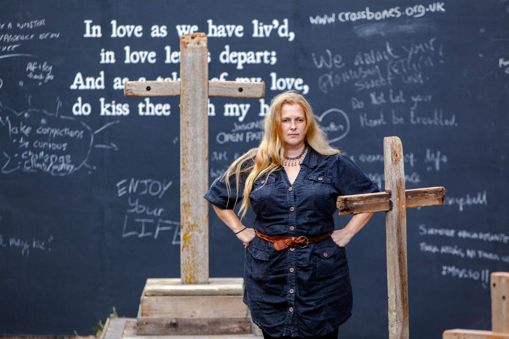 Emily Peasgood standing in front of her graveyard sound installation Requiem for Crossbones. There is a wooden cross behind her. She has her hands on her hips, and looks like she's thinking about something. It is a striking pose. 