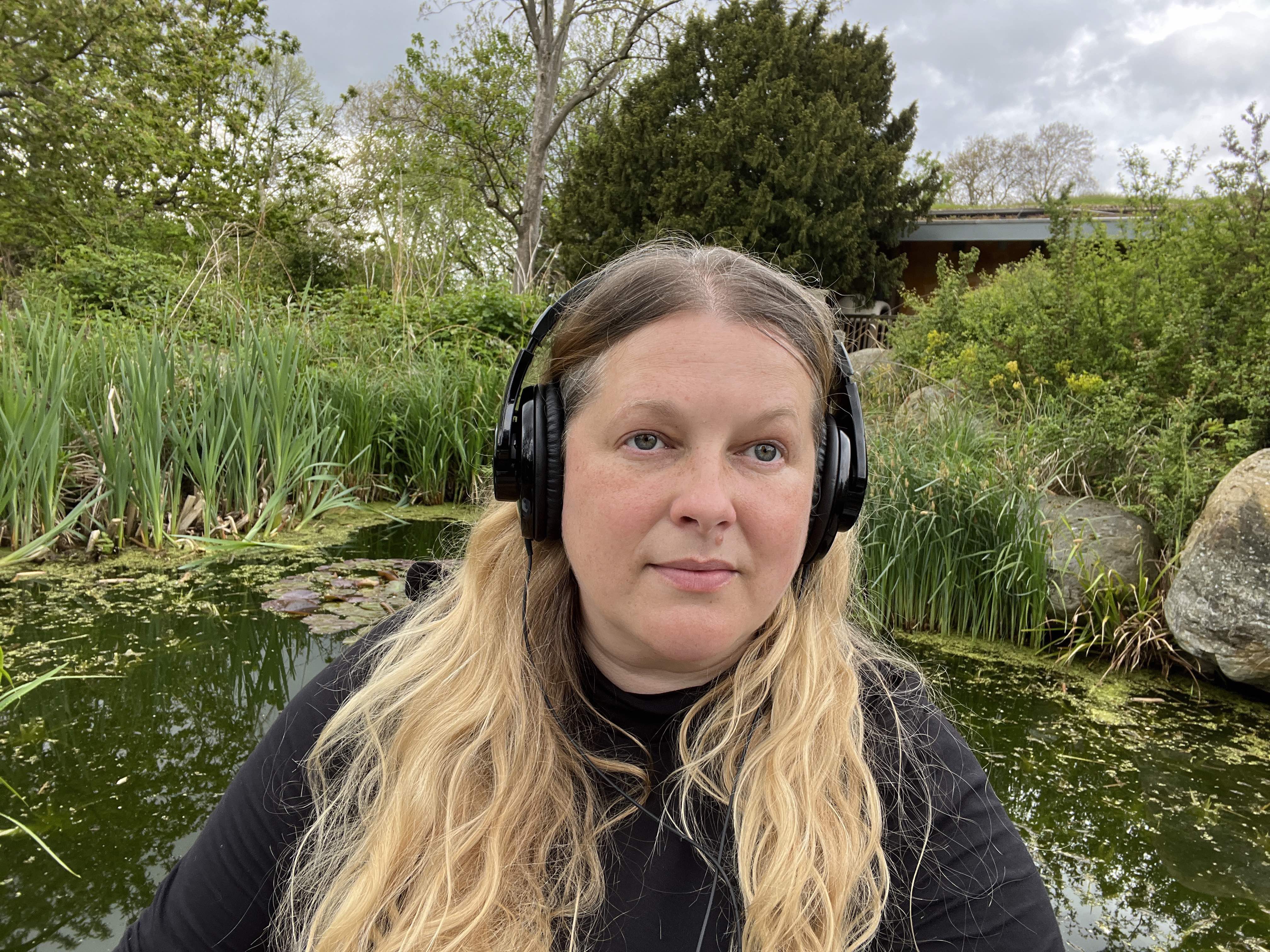 Emily Peasgood is standing in Hyde Park looking at the camera with a large pair of over-head earphones on. 